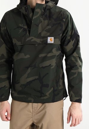 Mann trägt dunkelgrüne und schwarze camouflage Kapuzenjacke mit front zipper und beige Hose, steht vor einfarbigem Hintergrund.
