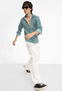 Gestreiftes türkis- und weißes Hemd mit Knöpfen, weite weiße Cargo-Hosen mit Reißverschlüssen, schwarze Sneakers und Sonnenbrille. Model in Bewegung.