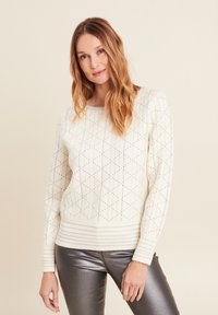Maglione crema con motivo geometrico, polsini e orlo a coste, con dettagli metallici. Indossato con pantaloni argentati lucidi per un contrasto.