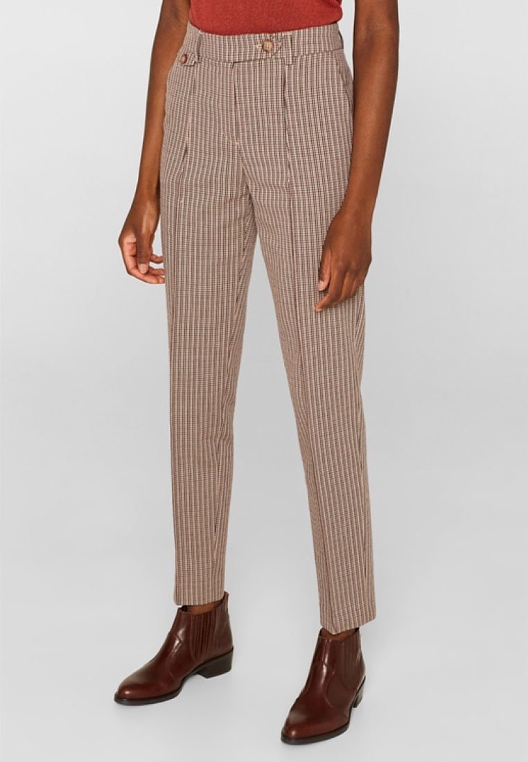 Femme portant un pantalon à carreaux beige et marron taille haute, un haut couleur rouille et des bottines en cuir marron, debout devant un fond uni.