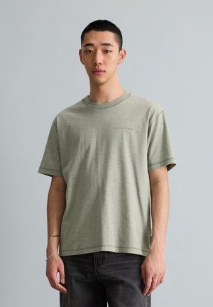 Jeune homme aux cheveux courts portant un t-shirt vert clair Marc O’Polo Denim et un pantalon foncé, debout devant un fond gris uni.