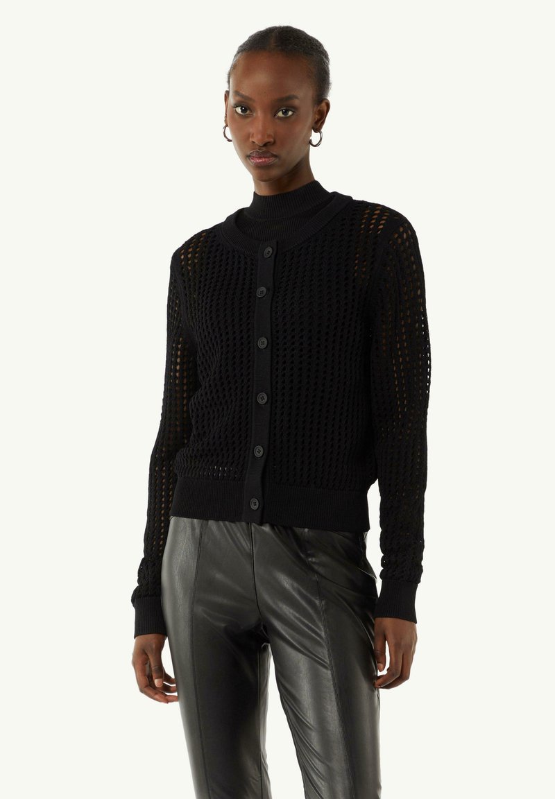 Zwarte gebreide cardigan met een gestructureerde open weving, een geribde kraag en manchet, en knoopsluiting. Gestyled met strakke zwarte leren broeken.