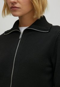 Schwarzer Reißverschluss-Pullover mit geripptem Kragen, strukturiertem Stoff und silbernem Reißverschluss. Nahaufnahme des Hals- und oberen Brustbereichs.
