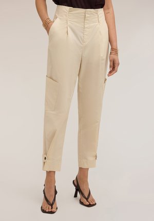 Beige broek met hoge taille, voorplooien, zijzakken, geknoopte enkelbandjes, gedragen met zwarte sandalen met hoge hakken en gouden armbanden.