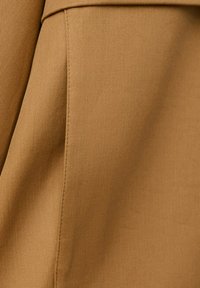 Gros plan sur un tissu beige avec une texture de tissage diagonal visible et une couture, montrant une partie d'un vêtement.