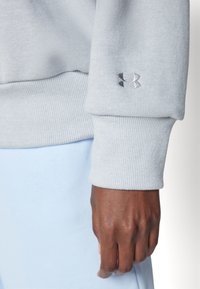 Detailný záber na tmavú ruku v svetlosivej mikine s kapucňou Under Armour a svetlomodrých nohaviciach.