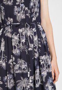 Robe bleu marine avec imprimé blanc de palmier tropical et d'animaux, avec un cordon noué à la taille et vue partielle du bras d'une personne.