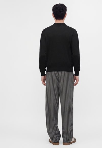 Uomo in piedi di spalle, indossa una maglia nera a maniche lunghe, pantaloni larghi a righe verticali grigie e scarpe marroni su uno sfondo semplice.
