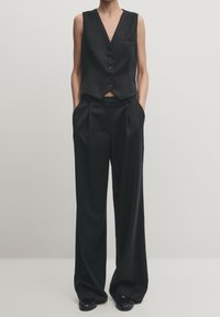 Czarna dopasowana kamizelka z głębokim dekoltem w kształcie litery V i pięcioma guzikami, w połączeniu z czarnymi spodniami o szerokich nogawkach z ostrymi plisami i kieszeniami.
