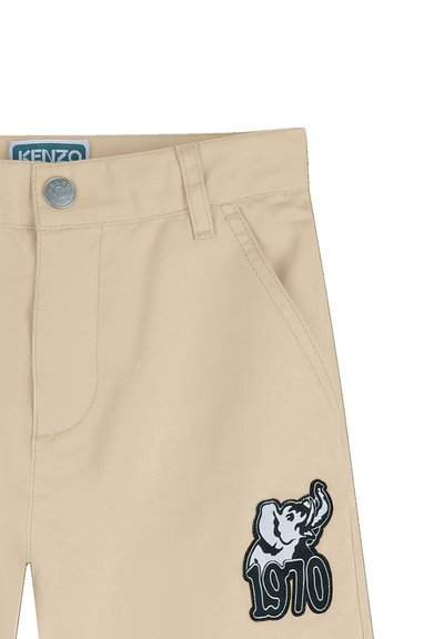 Pantalon beige avec bouton argenté, poches avant, passants pour ceinture, étiquette "KENZO" à la taille, et écusson éléphant noir et blanc avec "1970" sur la cuisse.