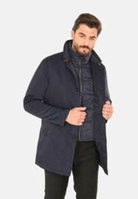 Cappotto blu navy con rivestimento interno trapuntato, chiusura a bottoni e colletto rialzato. Presenta una texture liscia e dettagli di cuciture a contrasto.