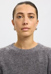 Siv pleten pulover z okrolim izrezom in teksturiranim tkanjem. Model nosi majhne srebrne obročaste uhane, z nevtralnim ozadjem.