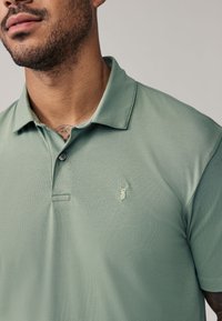 Mand iført en lysegrøn poloshirt med broderet hjortehovedlogo på brystet, der viser en del af sit skæg og en nakkedragt.