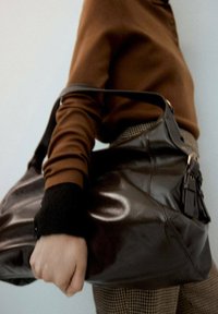 Hnedá kožená kabelka s lesklým povrchom, veľkého tvaru, nastaviteľným popruhom a leštenou kovovou bižutériou. Čiastočne sú viditeľné textúrované kárované nohavice.