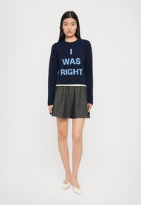 Femme debout portant un pull bleu marine avec le texte "J'AVAIS RAISON", une jupe plissée grise et des chaussures blanches à bouts pointus sur un fond uni.