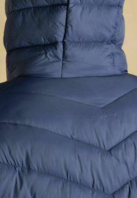 Marineblå pufferjakke med et chevron-quiltet mønster. Har en høj krave og det præget mærkenavn "JOULES" på ryggen. Glat tekstur.