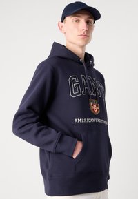 Sudadera azul marino con un logo central en blanco con detalles en rojo y amarillo. Incluye un bolsillo frontal y capucha ajustable.