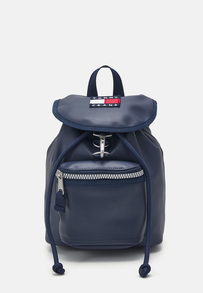 Sac à dos bleu marine avec un rabat arrondi, des sangles ajustables, une poche zippée à l'avant et un patch logo. Texture lisse, fabriqué en matériau durable.
