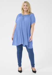Tunic azzurra chiara a maniche corte con scollo arricciato, tessuto fluido e orlo asimmetrico, abbinata a jeans skinny scuri e tacchi neri.