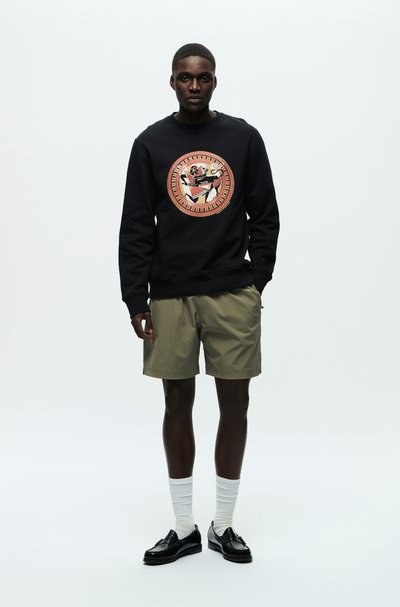 Svart sweatshirt med en central cirkulär grafik som föreställer en björn, tillsammans med olivgröna shorts. Modellen bär vita ribbade strumpor och svarta skor.