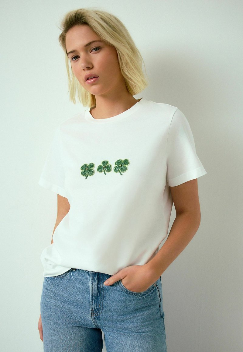 Giovane donna con capelli biondi indossa una maglietta bianca con tre trifogli verdi e jeans azzurri, in piedi contro una parete semplice.