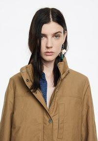 Giacca marrone con zip e colletto alto, bottoni a pressione e tasche laterali, indossata sopra una camicia a quadri blu. Tessuto liscio.