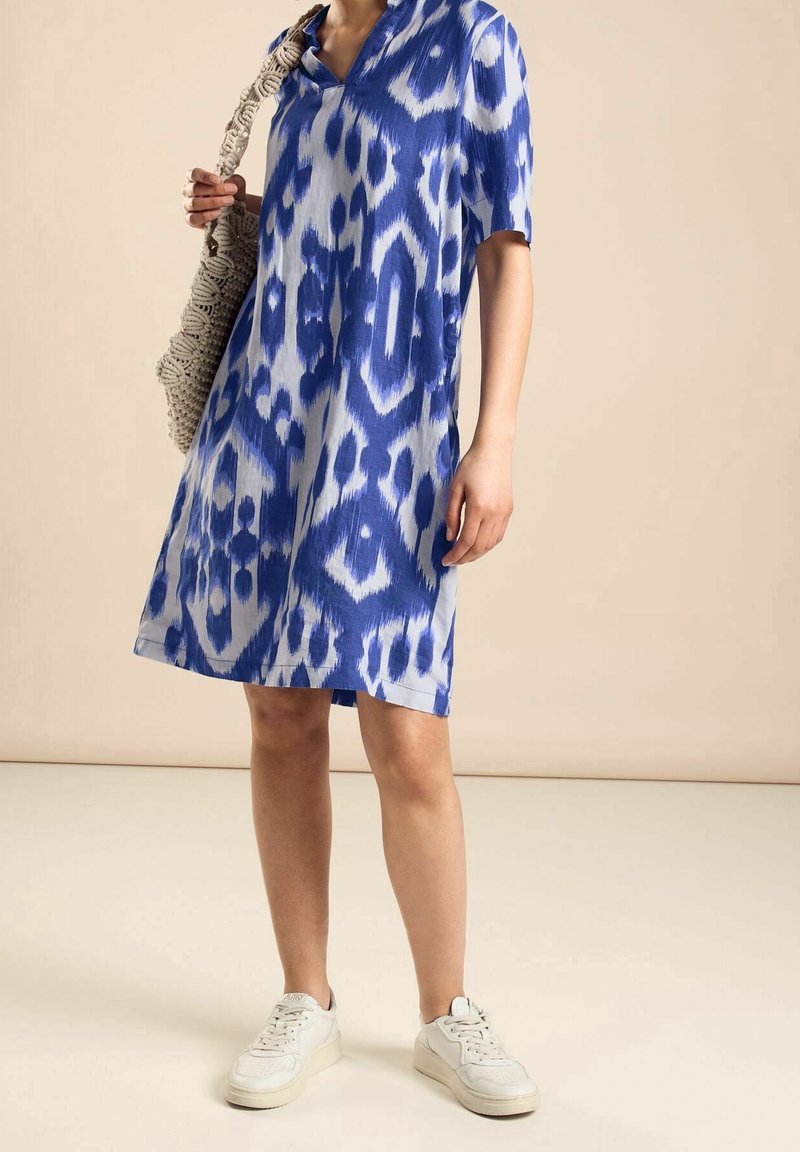Niebieska sukienka w wzory o luźnym kroju, z dekoltem w serek i krótkimi rękawami, sparowana z tkanym torbą i białymi trampkami. Gładka tekstura materiału.
