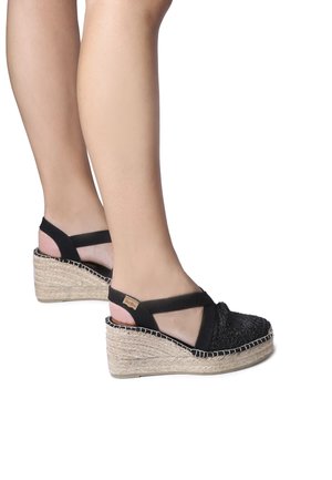 Black woven espadrille wedge sandals with elastic straps and a natural jute platform, featuring contrasting stitching along the edge.