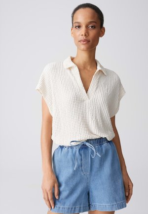 Donna che indossa una camicetta bianca a maniche corte con texture infilata in pantaloncini di jeans azzurri a vita alta con coulisse in vita.