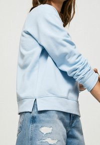 Persona che indossa un pullover a maniche lunghe di colore azzurro chiaro con polsini a coste e jeans in denim blu consumati, su uno sfondo neutro.