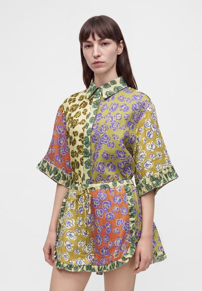Γυναίκα με πολύχρωμο φόρεμα patchwork με floral μοτίβα, κοντά μανίκια, γιακά και βολάν, στέκεται μπροστά σε απλό φωτεινό φόντο.