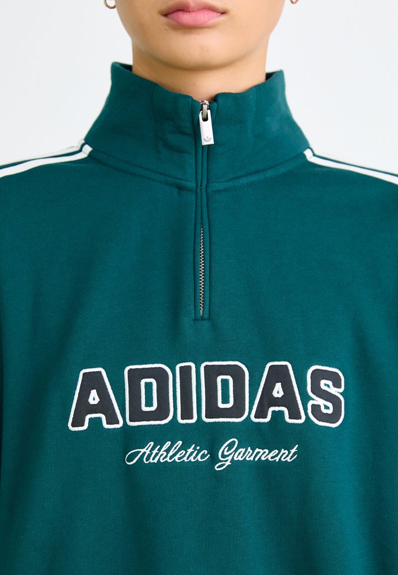 Sweatshirt zippé teal avec des rayures blanches sur les côtés, un col montant et "ADIDAS Athletic Garment" brodé en texte contrasté.