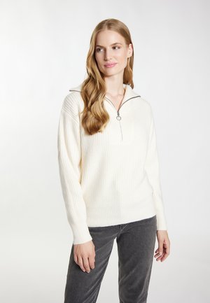 Donna con capelli lunghi e mossi indossa un maglione bianco lavorato a maglia con cerniera e pantaloni grigio scuro, in piedi su uno sfondo chiaro uniforme.