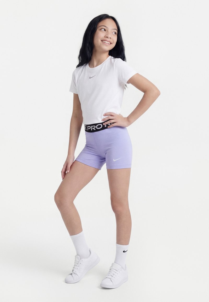 Κορίτσι με λευκό t-shirt Nike και λιλά σορτς Nike ποζάρει με το χέρι στη μέση μπροστά σε απλό λευκό φόντο.