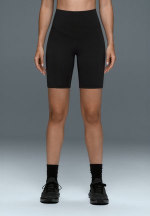 Personne portant un cuissard de cyclisme noir taille haute, des chaussettes noires et des chaussures de sport noires, debout avec les bras détendus le long du corps.