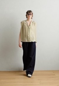 Lys olivgrønn ermeløs bluse med knappestolpe foran, sammen med løstsittende mørke denimbukser og hvite joggesko. Enkel design, uformell stil.