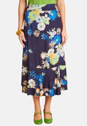 Frau trägt einen marineblauen Midirock mit Blumenmuster in Grün, Blau und Gelb, grünes Oberteil, goldene Armreifen und grüne Mary Jane Schuhe.