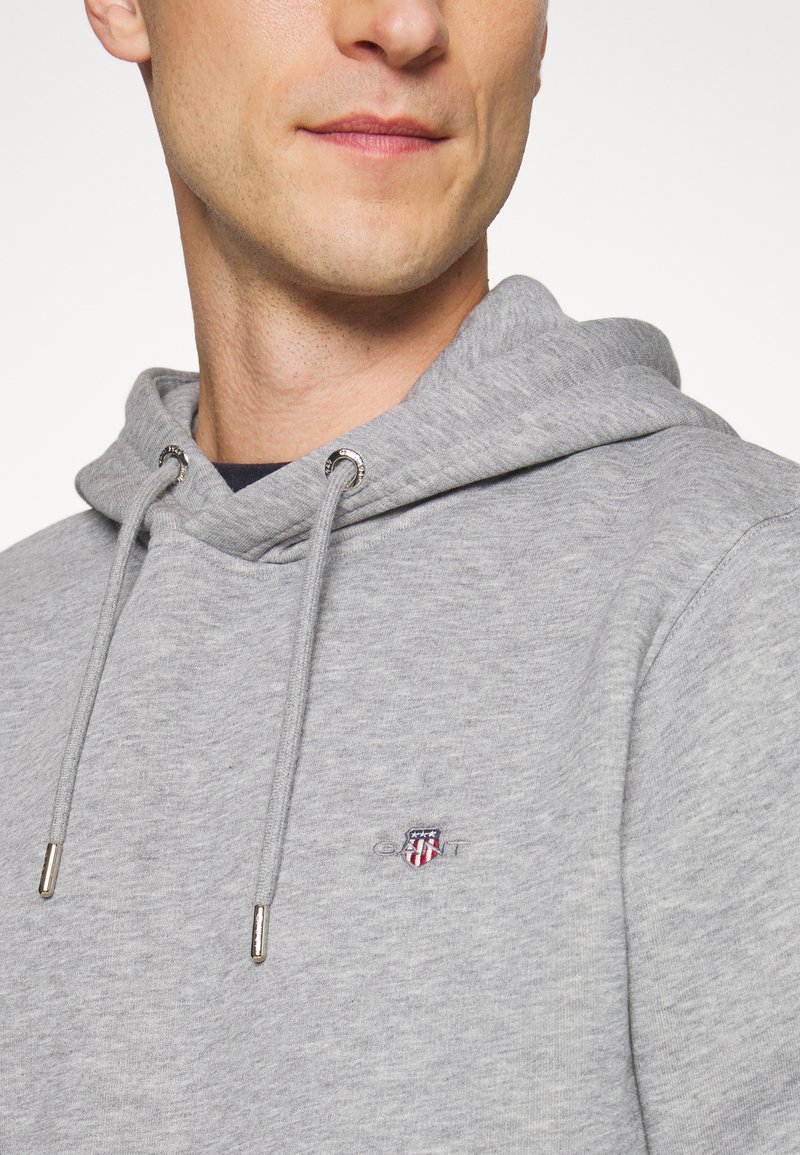 Hombre con sudadera con capucha gris claro, con cordones en la capucha y pequeño logo bordado en escudo rojo, blanco y azul en el pecho.