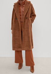 Abrigo de pelo sintético marrón con un diseño de doble botonadura, grandes solapas y bolsillos laterales, superpuesto a una chaqueta acolchada a juego y a pantalones.