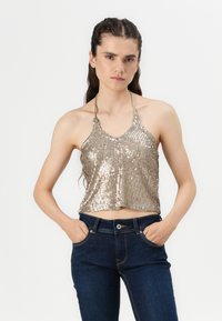 Zlati bleščičasti crop top z halter ovratnikom in valovitimi robovi. V kombinaciji s temno modrimi kavbojkami, ki imajo pet žepov in prilegajoč stil.