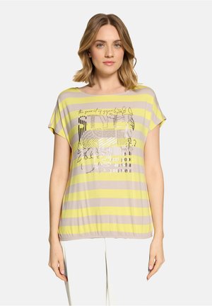 Vrouw draagt een geel-beige top met horizontale strepen, bloemenmotief en tekst, gecombineerd met een witte broek, staand in een neutrale houding.