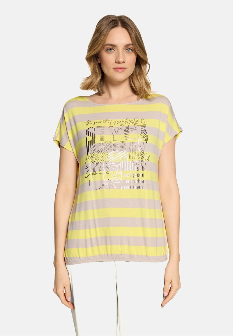 Femme portant un haut à manches courtes rayé horizontal jaune et beige avec des motifs floraux et du texte, associé à un pantalon blanc, en position neutre.