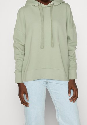 Person trägt einen hellgrünen, übergroßen Hoodie mit Kordelzug und hellblaue Jeans und steht vor einem schlichten Hintergrund.