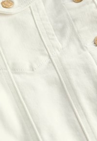 Fehér denim szövet, látható varrással, két mellzsebbel és kerek arany gombokkal. Sima tapintású, strukturált fazon.