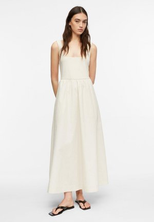 Młoda kobieta stojąca, ubrana w bezrękawnikową, kremową sukienkę maxi oraz czarne, płaskie sandały na jednolitym białym tle.