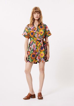 Robe courte à fleurs présentant des couleurs vives, des manches courtes, un devant à boutons et une ceinture à nouer à la taille. Portée avec des sandales à talons bruns.