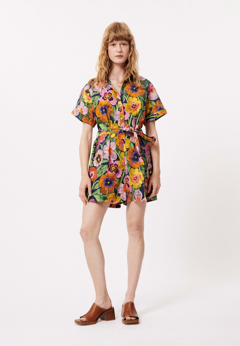 Robe courte à manches courtes à motifs floraux avec des couleurs vives, une taille ceinturée et des détails à boutons. Portée avec des talons blocs ouverts marron.