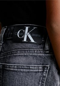 Jeans de mezclilla negra con una etiqueta visible que dice "Calvin Klein Jeans", que presenta un audaz logo CK en blanco y costuras en el bolsillo trasero.