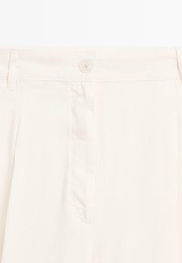 Pantalons beiges avec une texture de tissu lisse, fermeture à bouton à l'avant et poches latérales. Présente une taille cintrée et de légers plis.