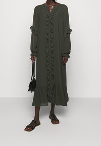 Vestido verde oscuro de manga larga con volantes en la parte delantera, los puños y el dobladillo. Combinado con sandalias negras y un pequeño bolso negro, sobre un fondo simple.